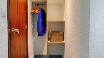 Un vestidor pequeño con puerta de madera, una chaqueta azul colgada en un perchero y una maleta de mimbre sobre una estantería.