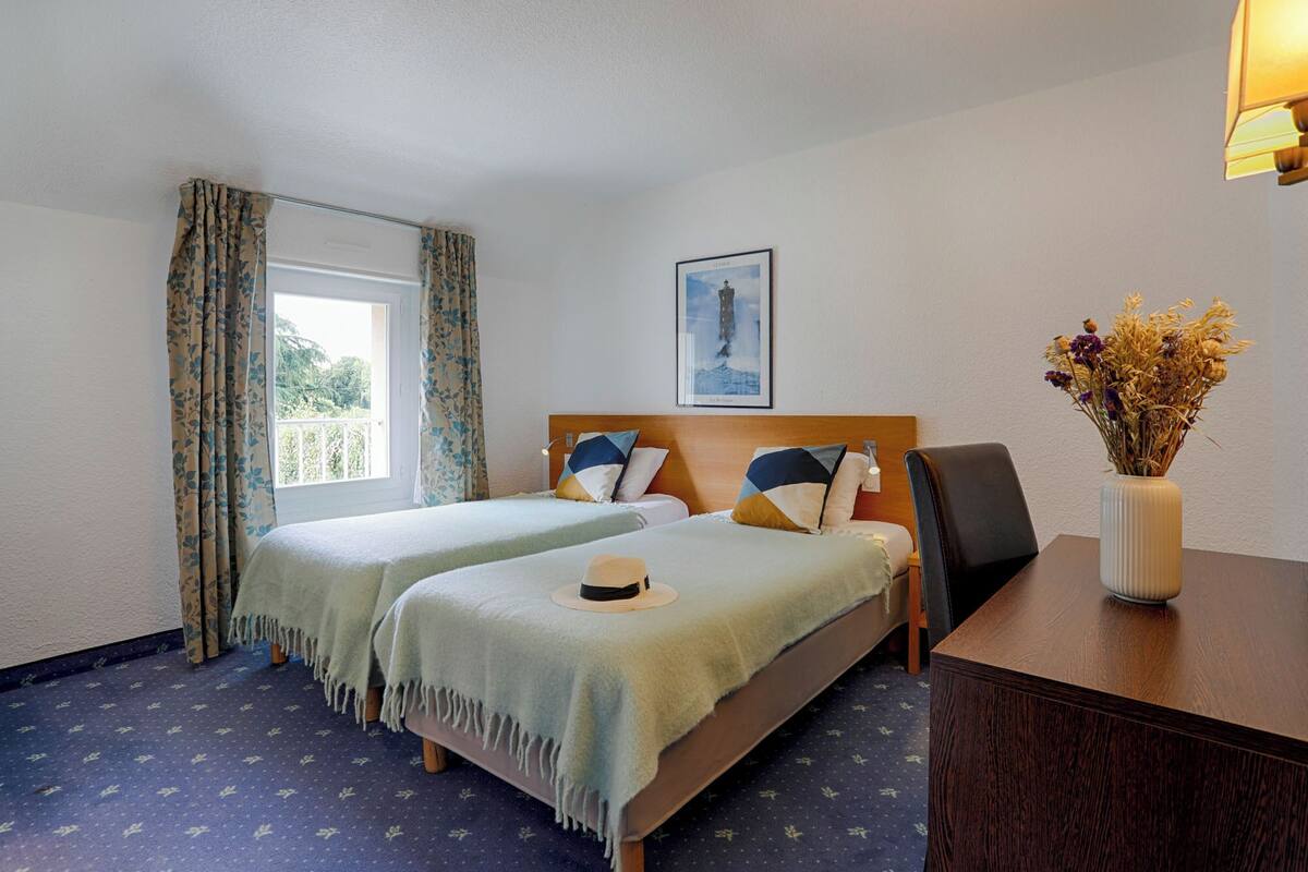 A hotel room with two beds, a wooden headboard, a desk with a vase of flowers, and a window with curtains.