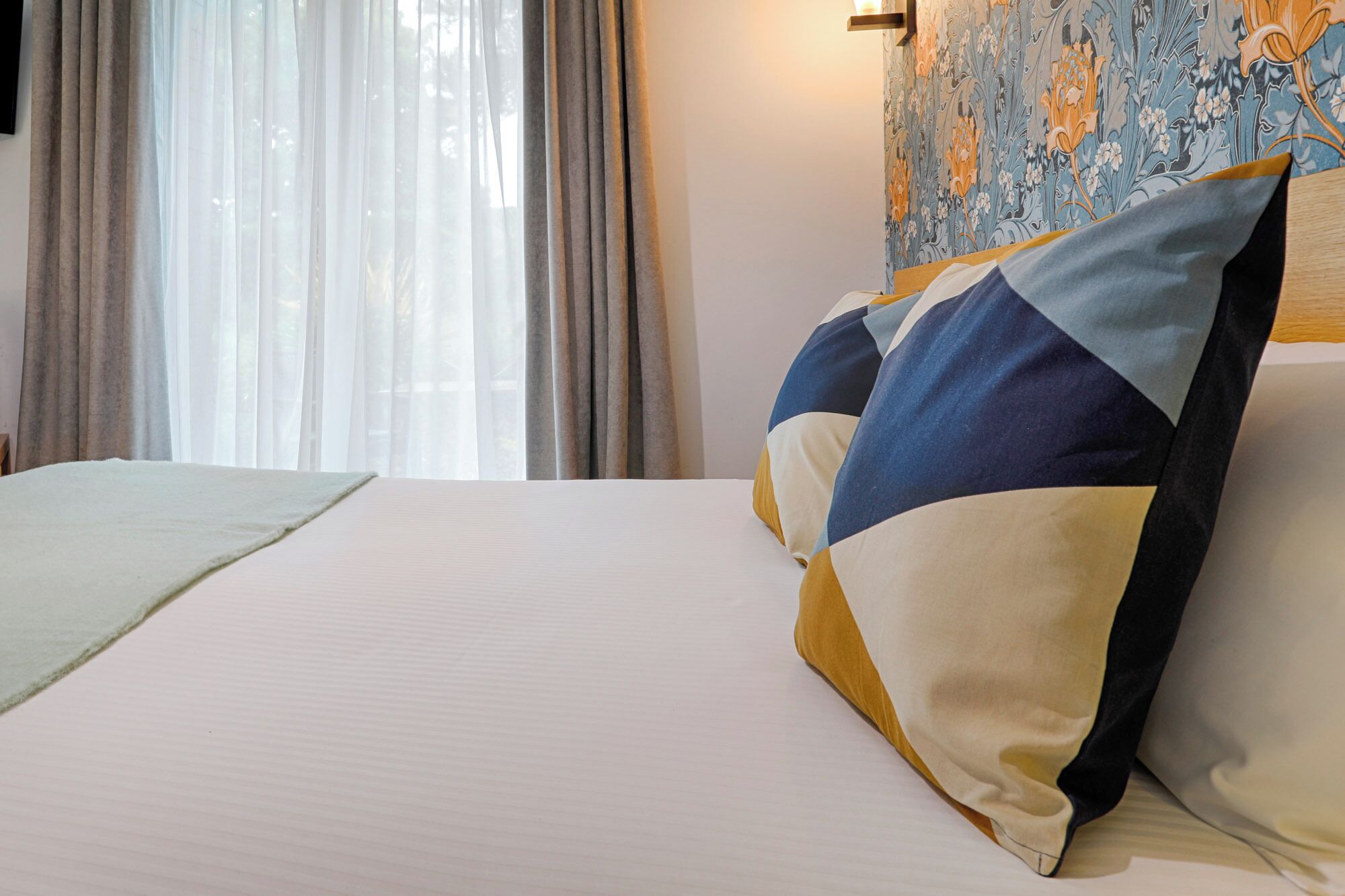Una habitación de hotel con una cama, un ventanal con cortinas traslúcidas y un papel tapiz con motivos florales.