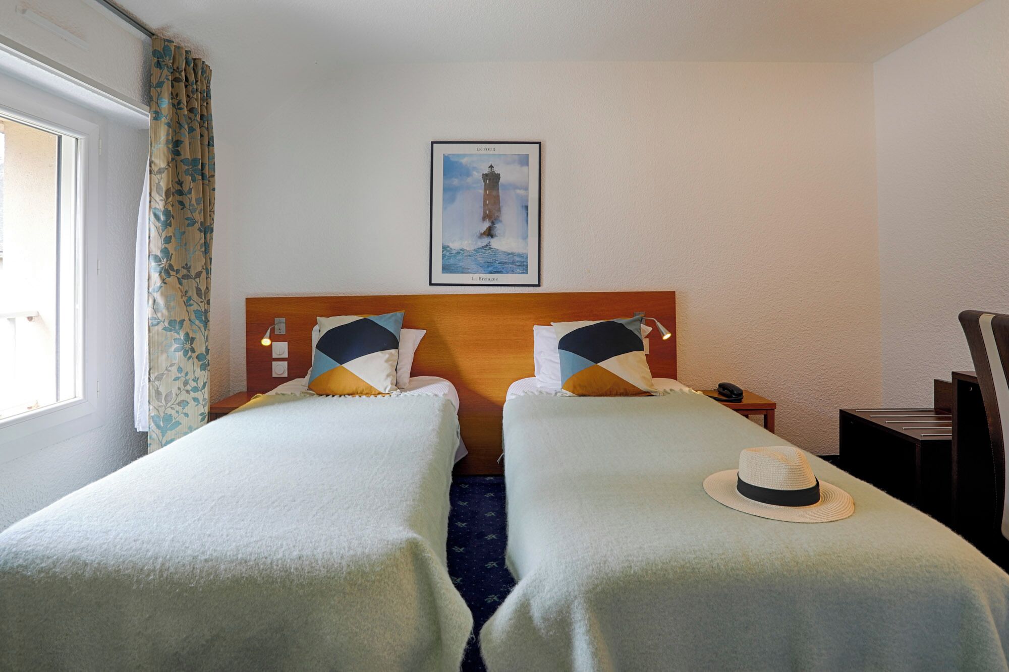 A hotel room with two beds, a wooden headboard, a framed picture of a lighthouse, a nightstand, and a desk with a telephone.