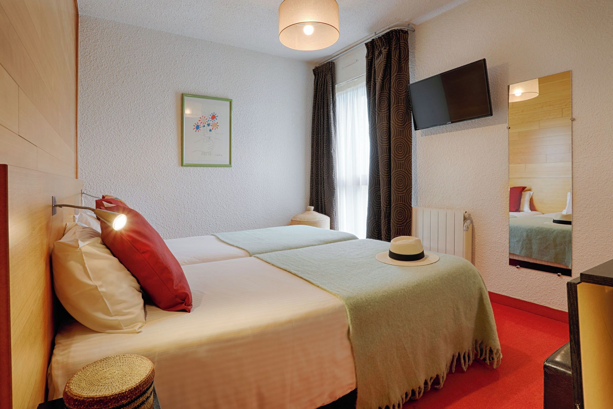 A hotel room with a bed, a bedside lamp, a small round table, a mirror, a television, and a framed picture on the wall.
