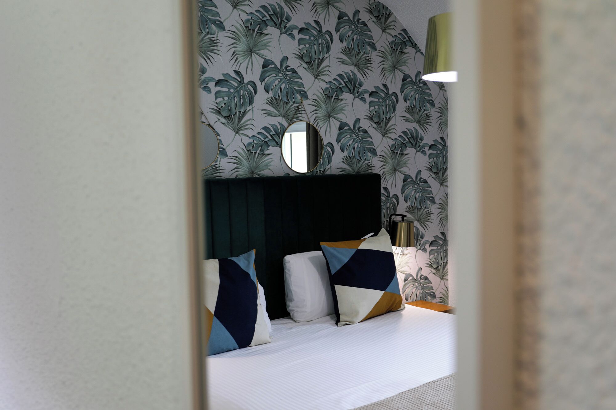 Un dormitorio con cama, cabecera oscura y pared con estampado de hojas tropicales.