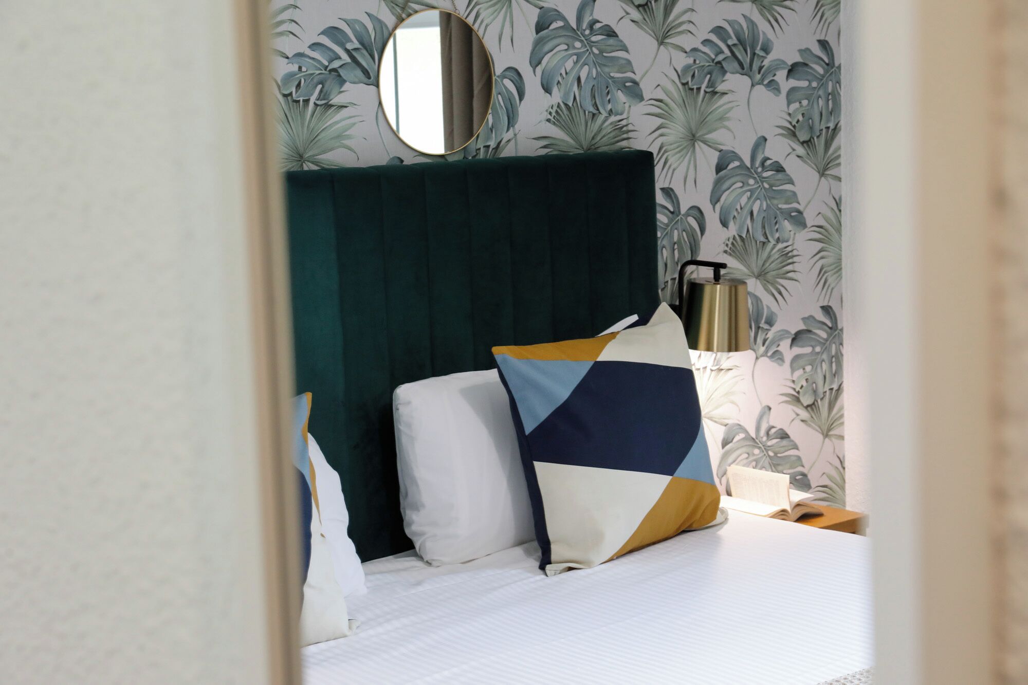 Ein Hotelzimmer mit Bett, Kissen, einem Nachttisch mit Lampe und einem Spiegel an der Wand.