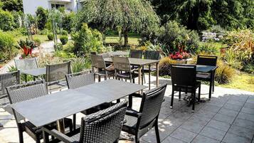 Un espace extérieur aménagé en terrasse, avec des tables et des chaises, entouré d’une végétation luxuriante et d’arbres.