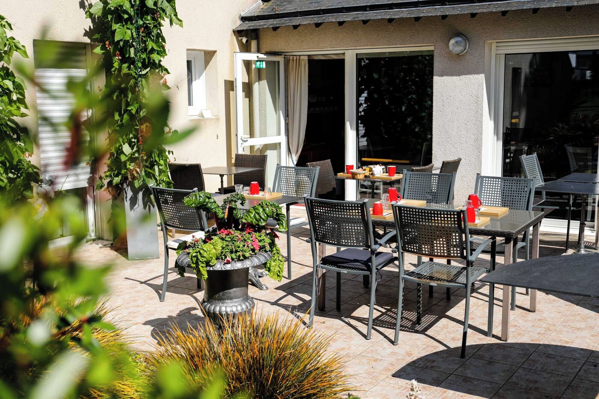 An outdoor patio area with multiple tables and chairs, surrounded by potted plants and a building with large windows.
