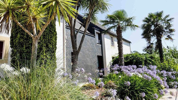 A modern house with a brick facade, surrounded by lush greenery and flowering plants. - Brit Hotel Le Branhoc (Auray)