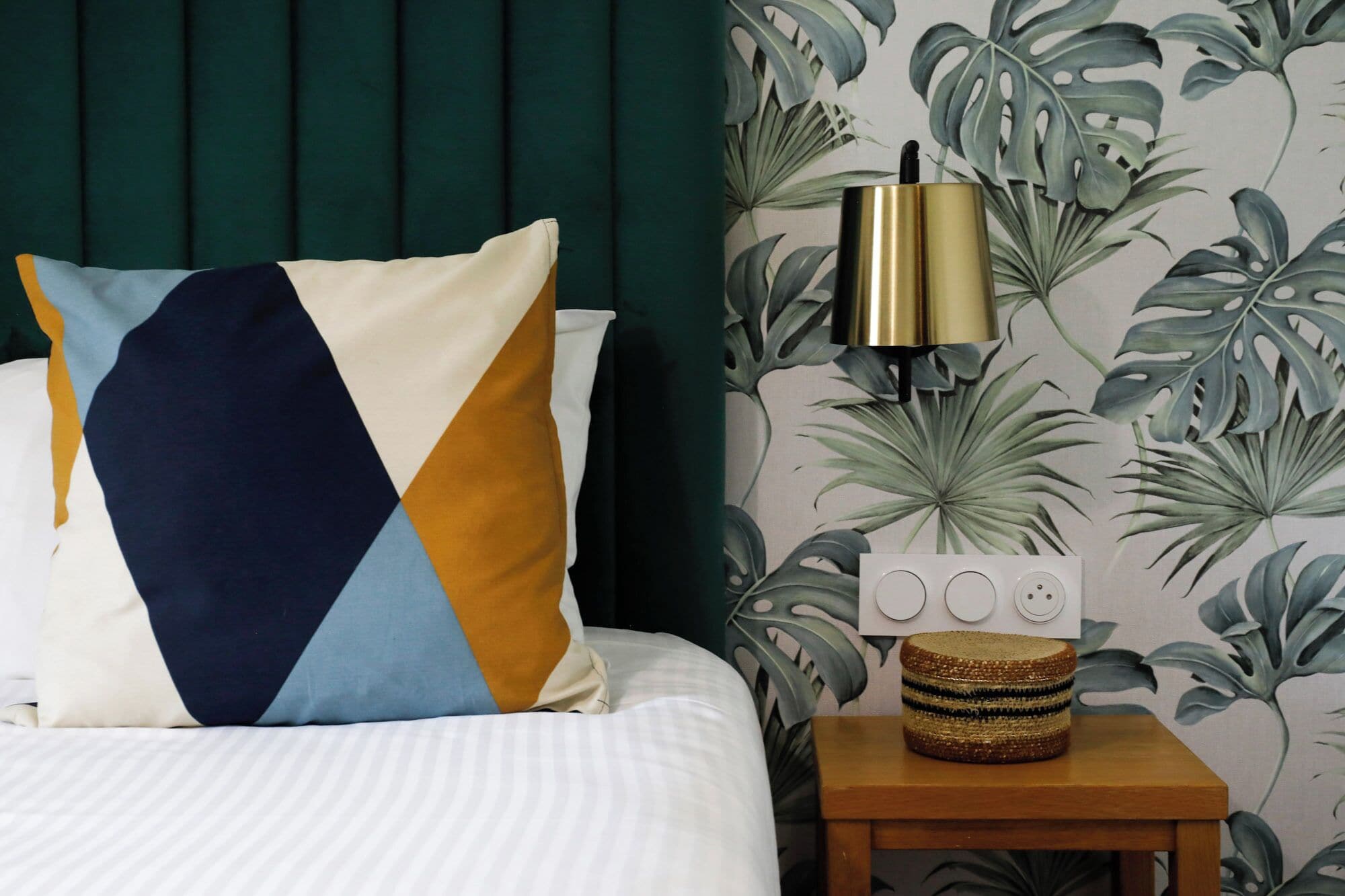 A neatly made bed with a geometric-patterned pillow, a bedside table with a lamp, and a wall with tropical wallpaper.