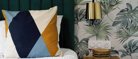 A neatly made bed with a geometric-patterned pillow, a bedside table with a lamp, and a wall with tropical wallpaper.