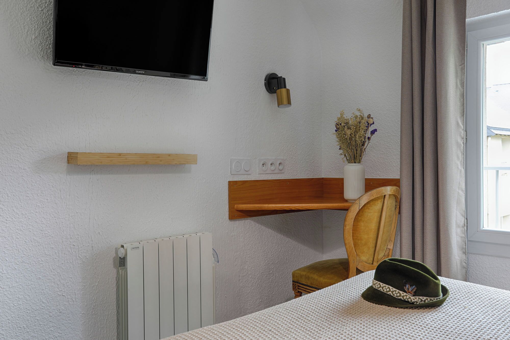 Ett hotellrum med en väggmonterad tv, en väggmonterad lampa, en hylla i trä, ett element, en stol, ett bord med en hatt på och ett fönster med gardiner.