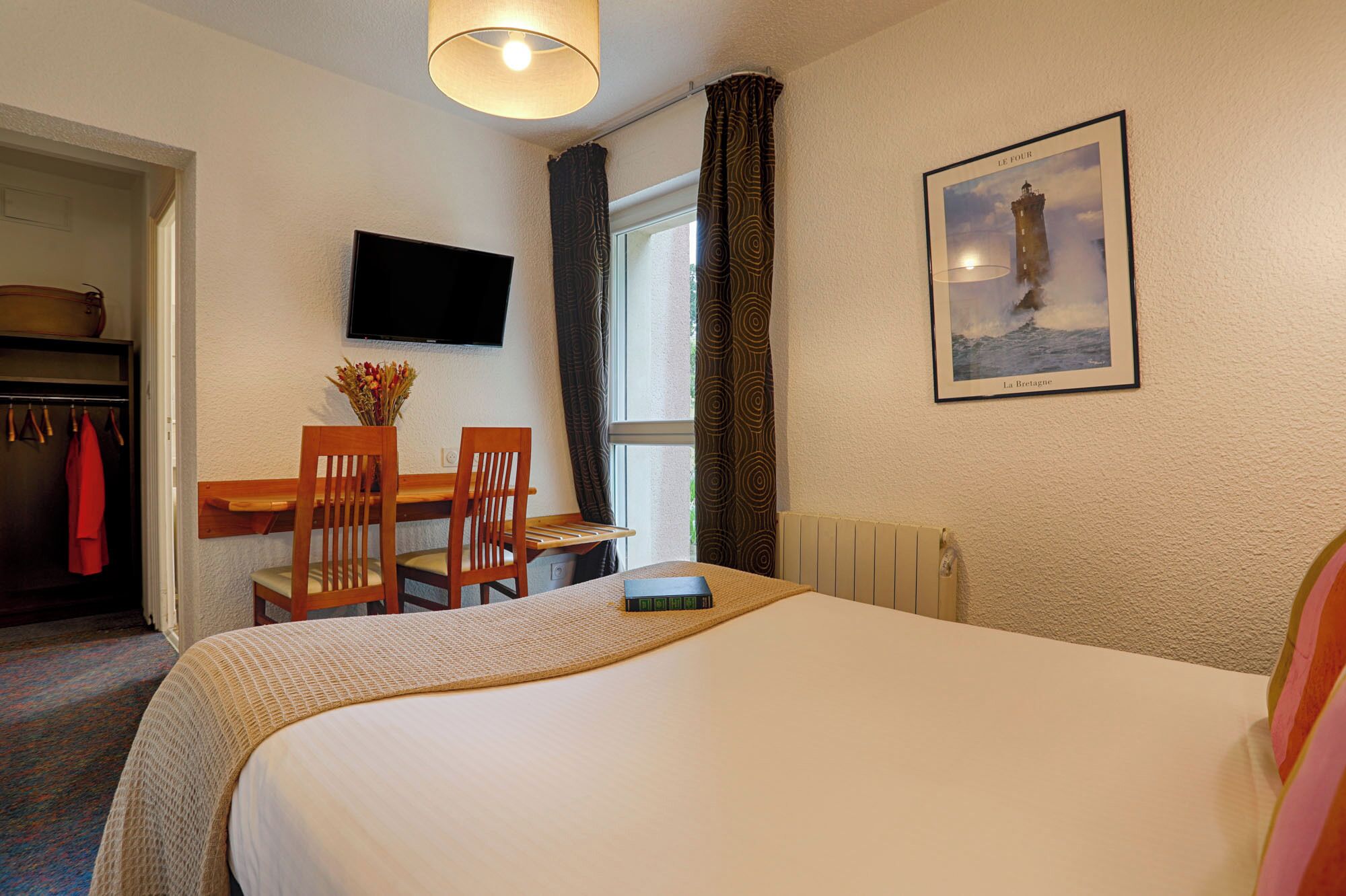 A hotel room with a bed, a desk with a chair, a TV, and a window with curtains.
