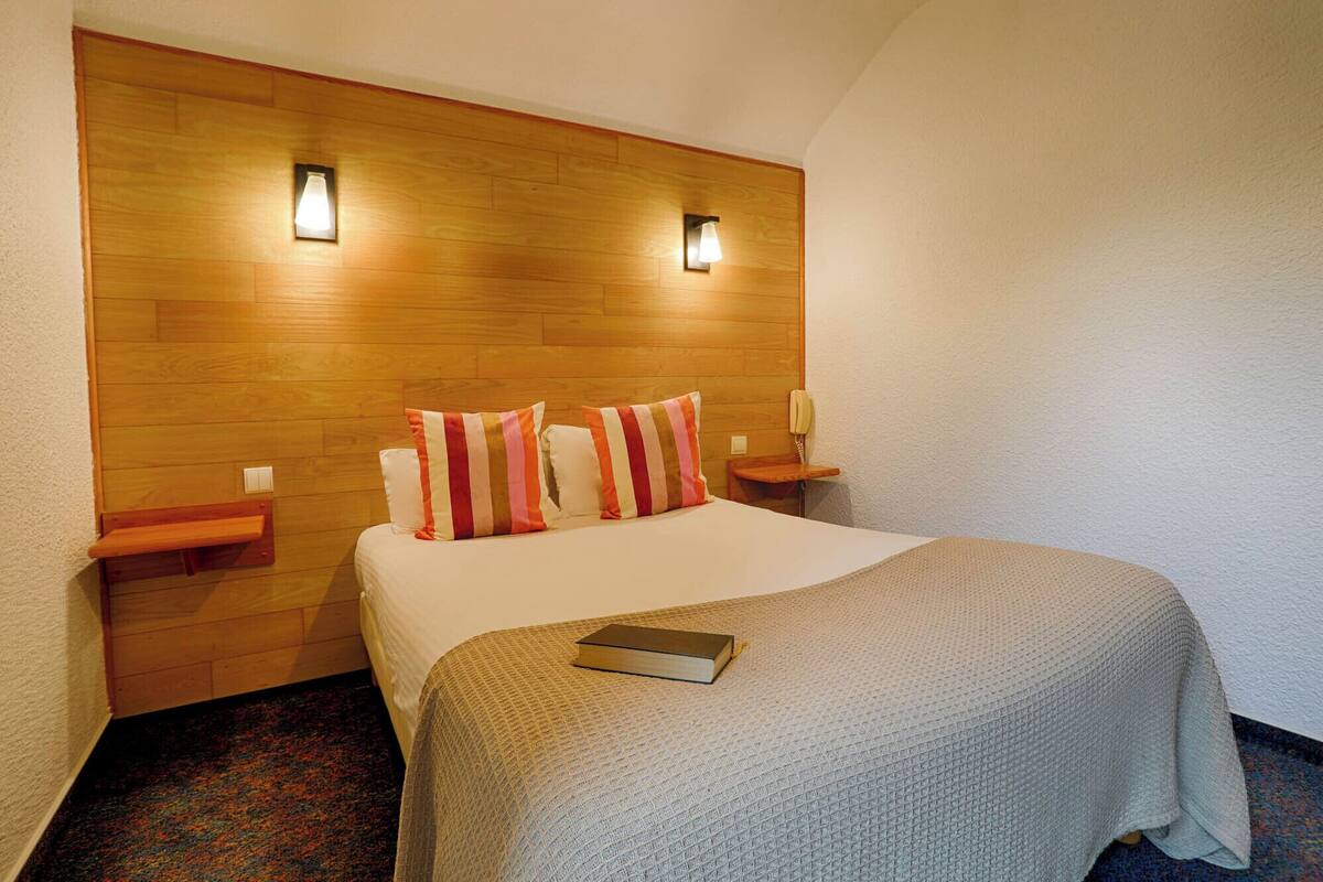 A hotel room with a wooden headboard, two bedside tables, a bed with a striped pillow, a bedside lamp, and a book on the bed.