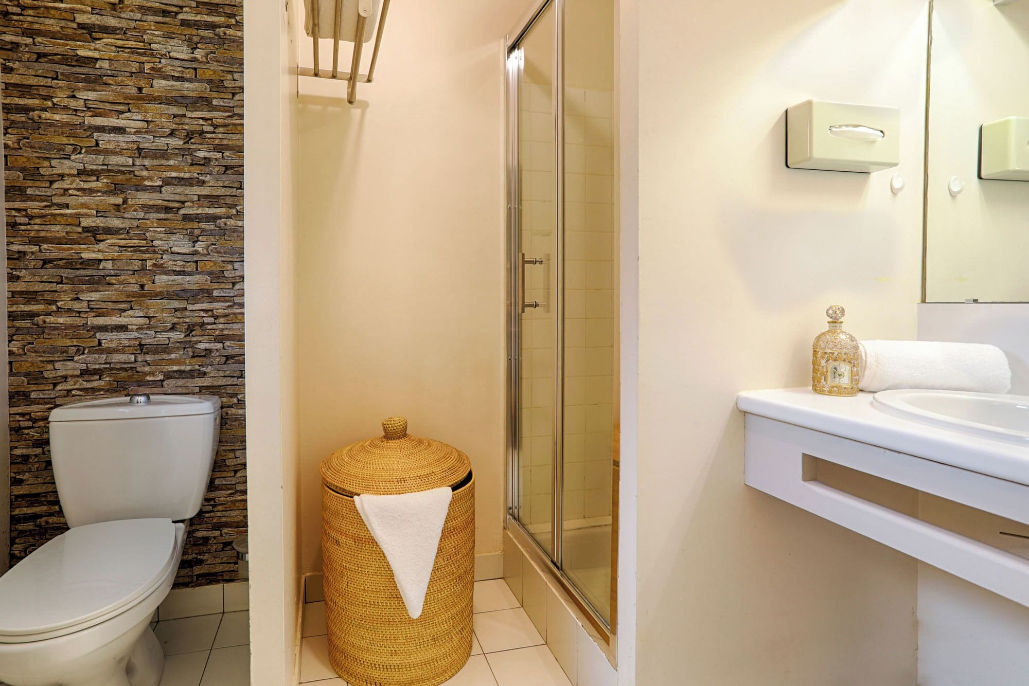 A bathroom with a stone wall, a toilet, a wicker trash bin, a glass shower enclosure, and a sink with a gold bottle.