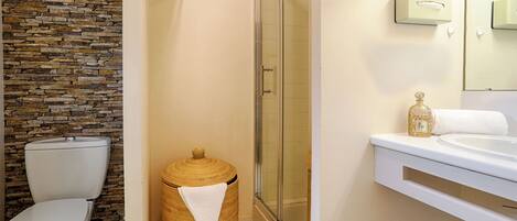 A bathroom with a stone wall, a toilet, a wicker trash bin, a glass shower enclosure, and a sink with a gold bottle.