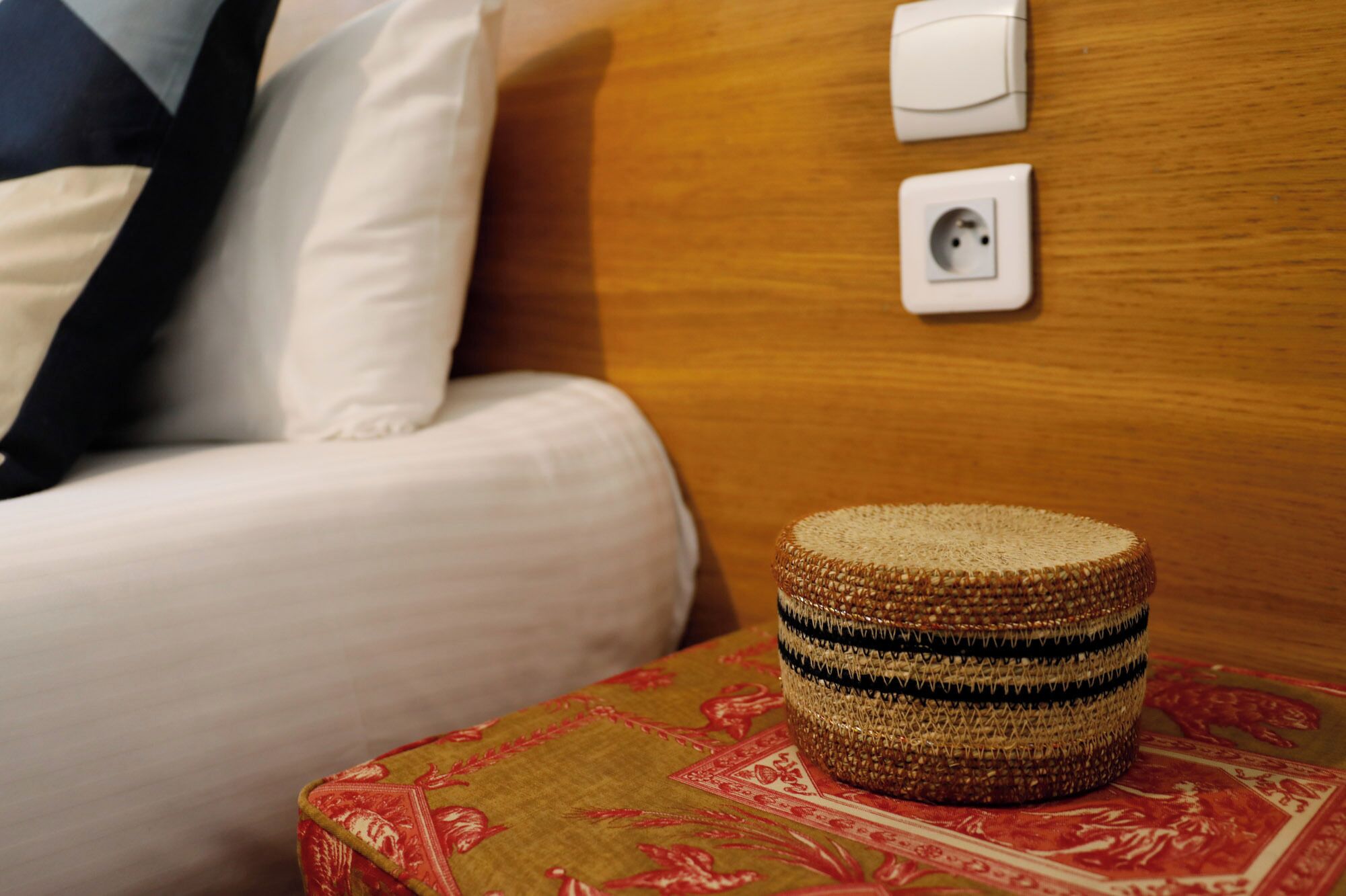 A bed with white linens and a wooden headboard, a power outlet, and a woven basket on a patterned cushion.
