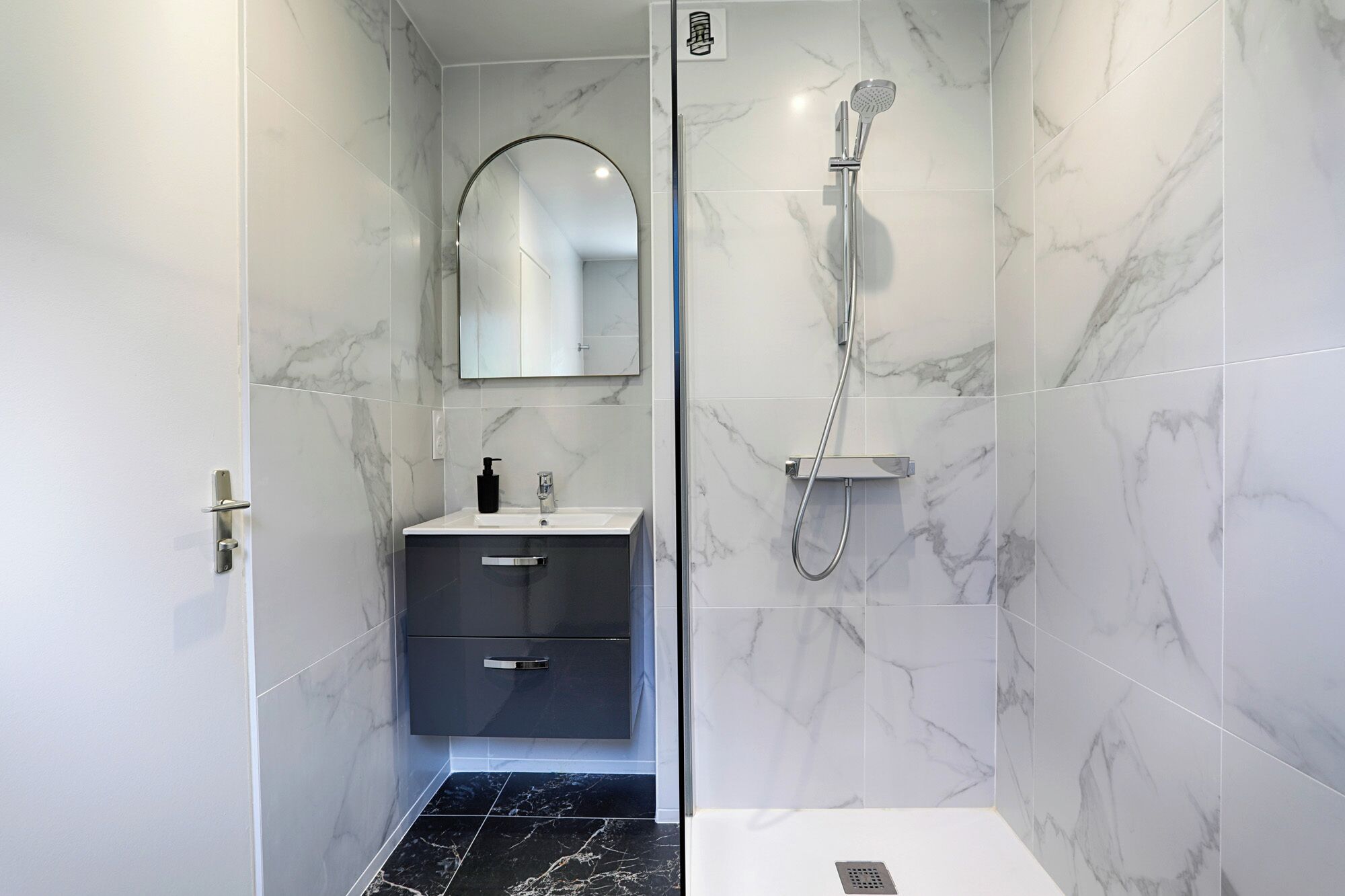Un baño moderno con baldosas de mármol, ducha, lavabo con espejo y suelo de mármol oscuro.