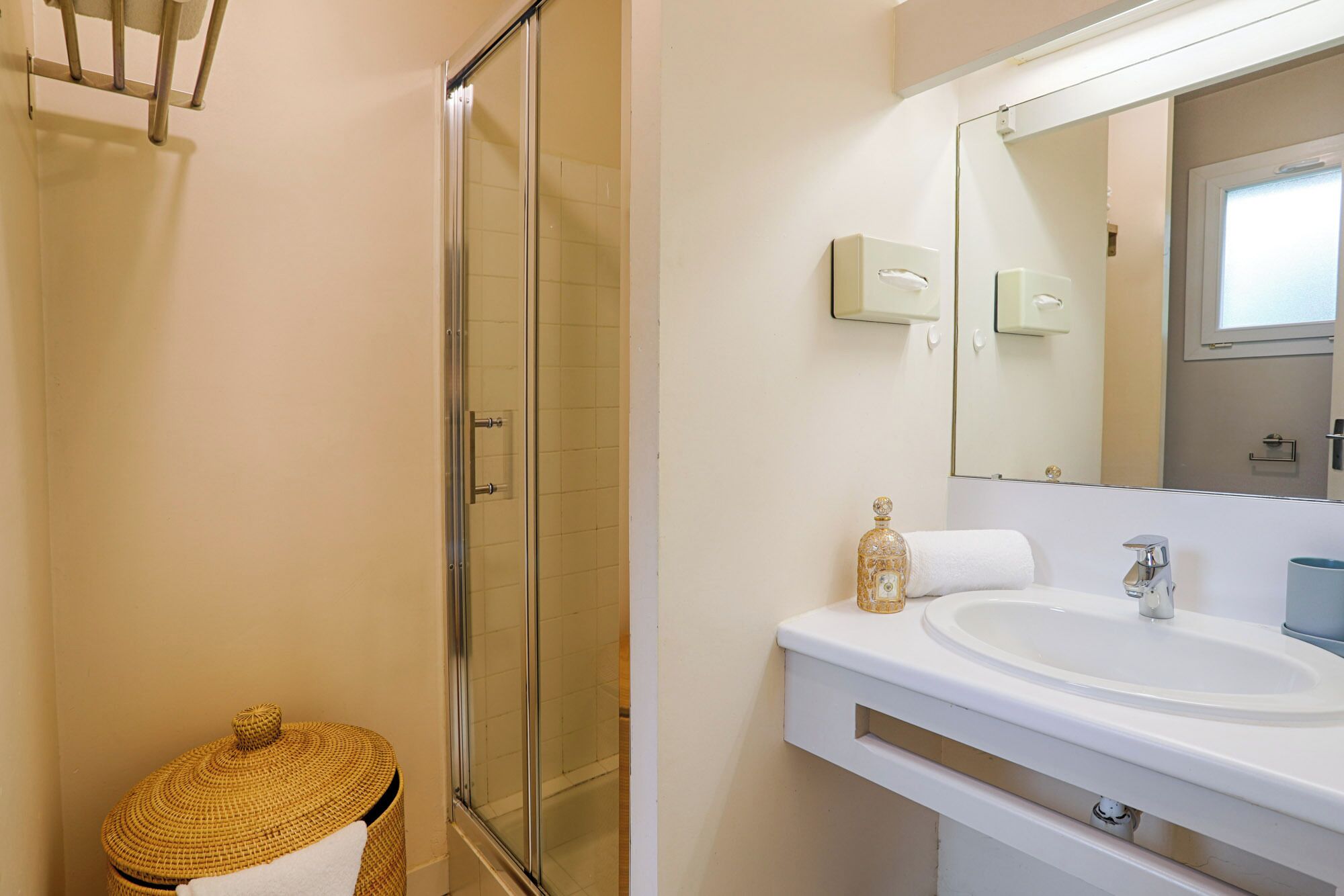 Une salle de bain avec une douche à parois de verre, un lavabo blanc, un grand miroir et un panier en osier.