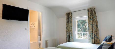 A hotel room with two beds, a TV mounted on the wall, a window with curtains, and a view of trees outside.