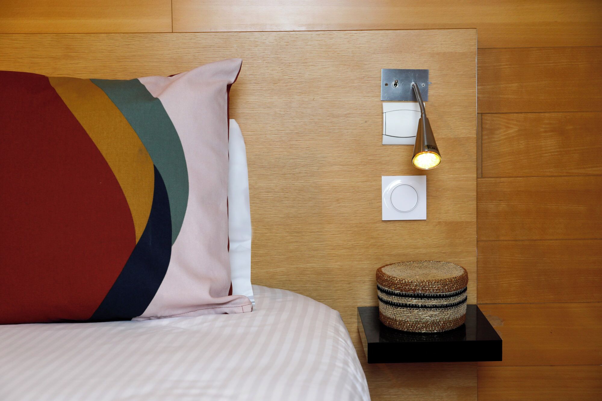 Een netjes opgemaakt bed met streepjesbeddengoed, een kleurrijk kussen, een bedlampje en een geweven mandje op een zwarte plank.