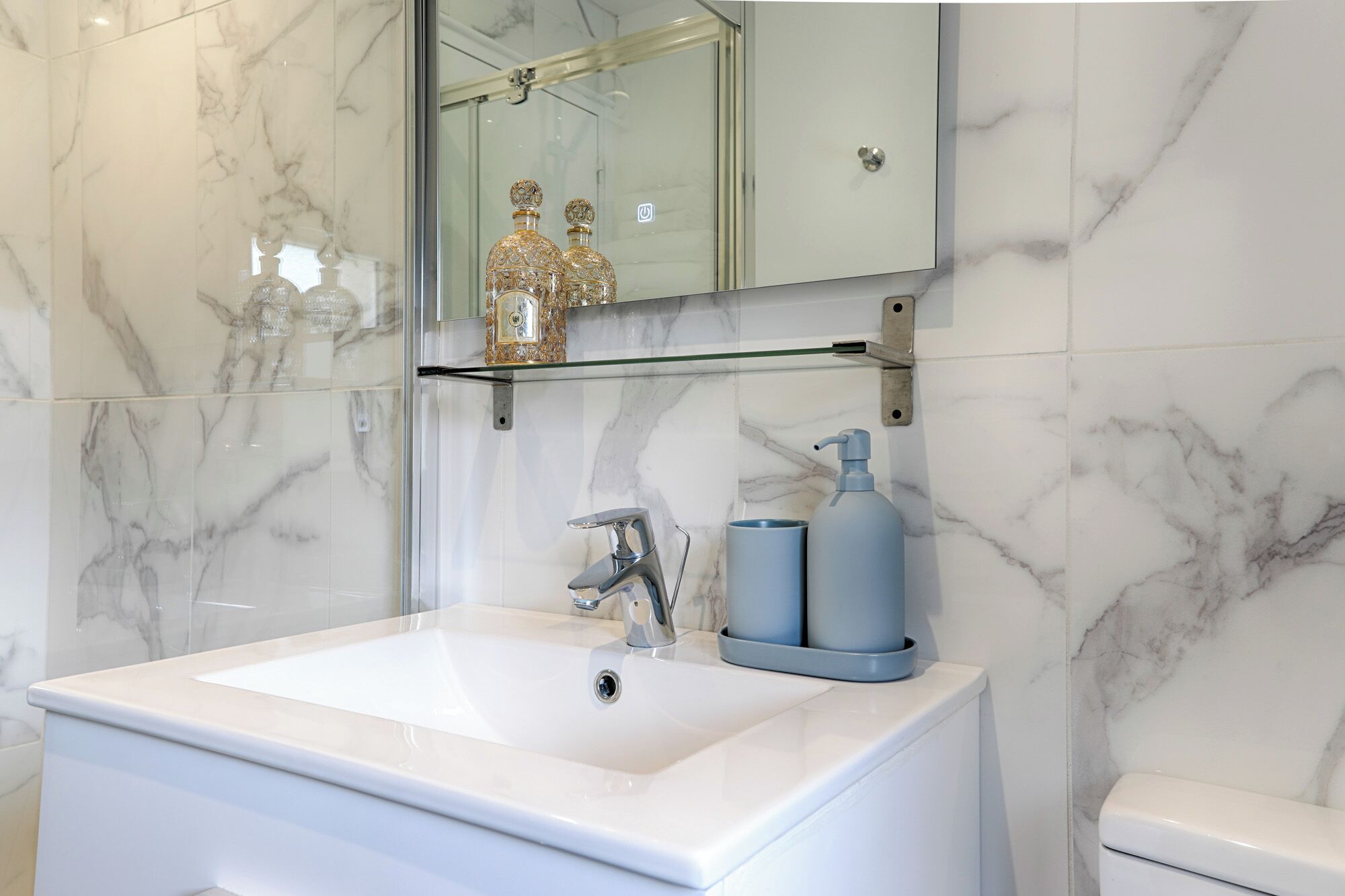 Une salle de bain avec un grand lavabo blanc, un miroir au-dessus et une étagère en verre où sont disposés des objets décoratifs.