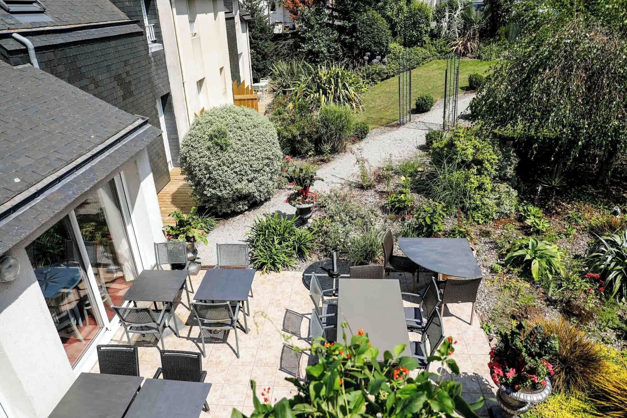 Un patio exterior con varias mesas y sillas, rodeado de plantas y un camino que lleva a un edificio.