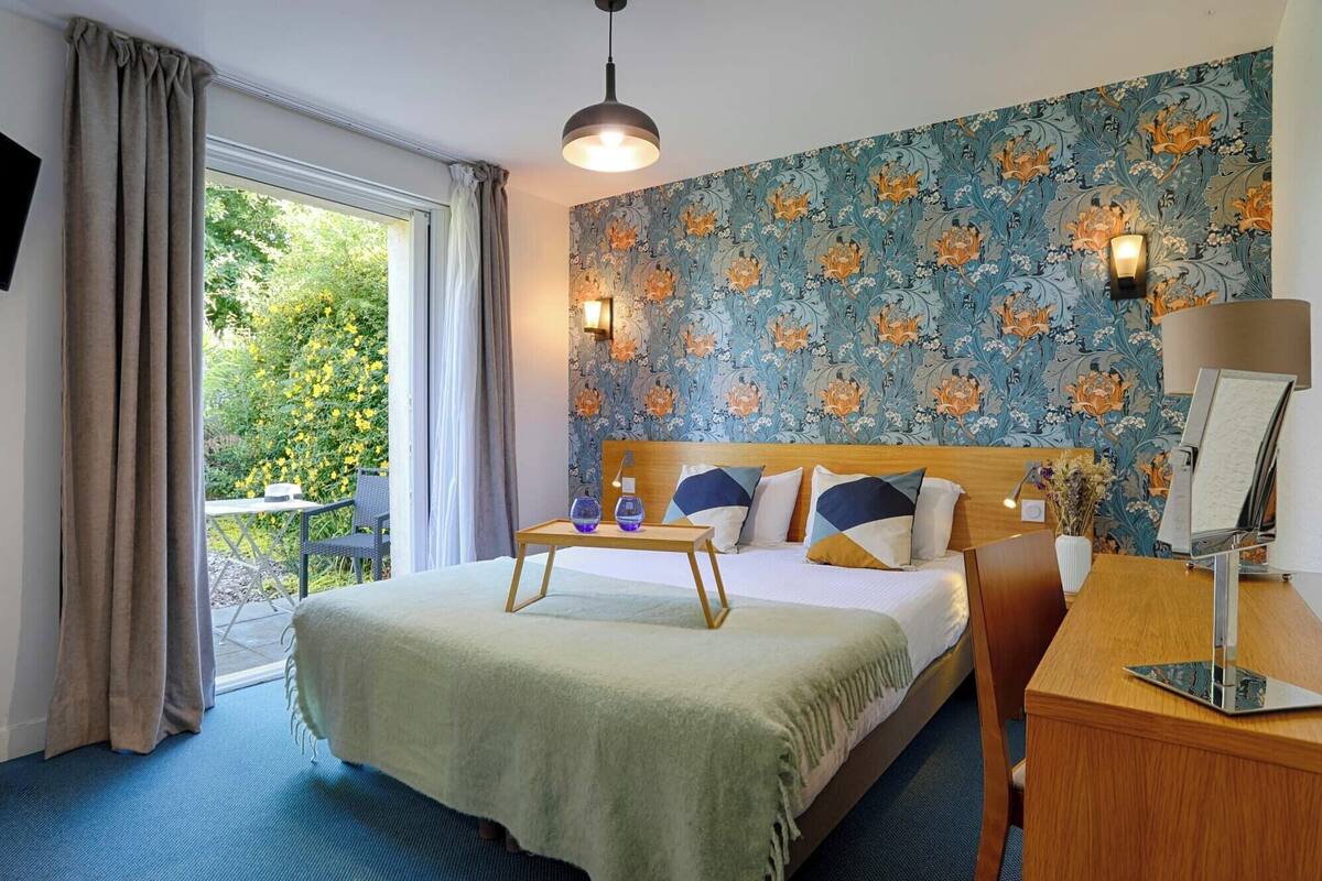 A hotel room with a bed, a desk, a chair, a lamp, and a view of greenery outside.