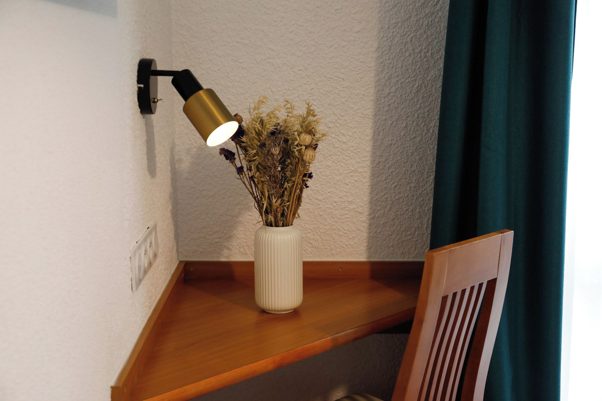 A corner desk with a white vase containing dried flowers, a black and gold desk lamp, and a wooden chair.