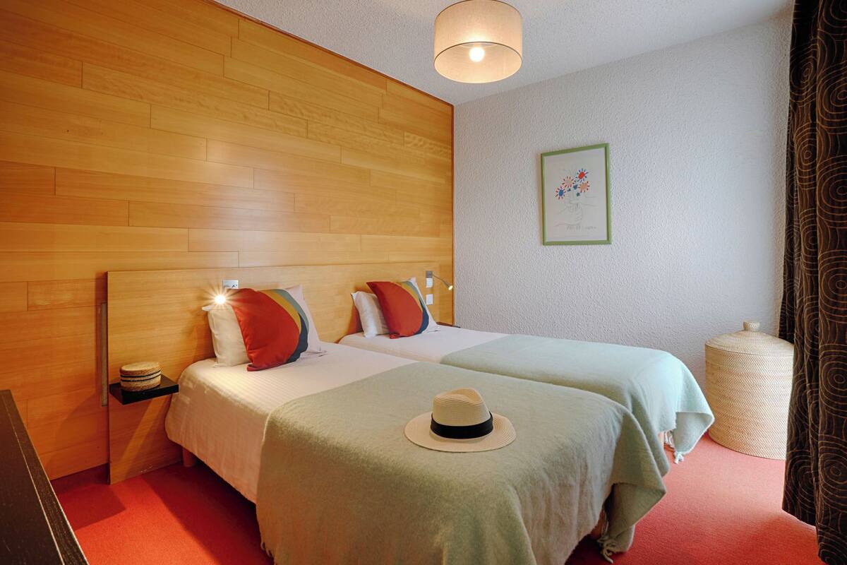 A hotel room with two beds, wooden headboards, a small nightstand, a lamp, a framed picture on the wall, and a woven basket.