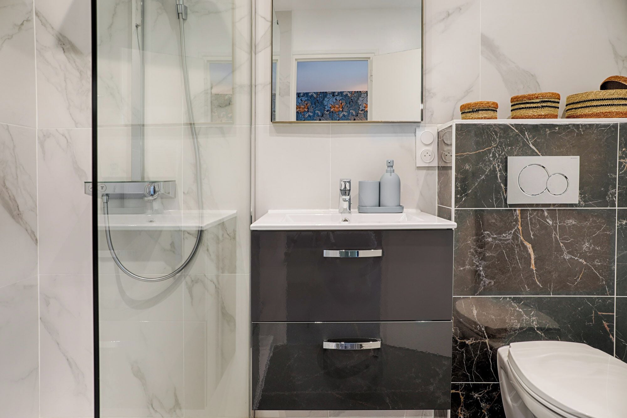 Un baño moderno con cabina de ducha de vidrio, un lavamanos blanco con grifo gris, un mueble negro con cajones, un inodoro y cestas decorativas en la pared.