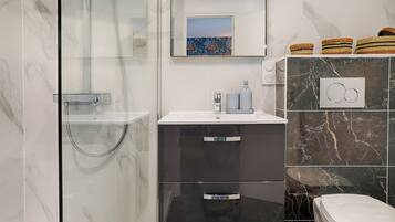 Un baño moderno con cabina de ducha de vidrio, un lavamanos blanco con grifo gris, un mueble negro con cajones, un inodoro y cestas decorativas en la pared.