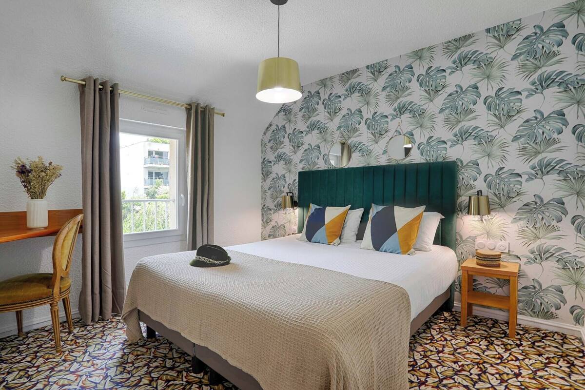 A hotel room with a bed, a desk, a chair, a window with curtains, a wall with tropical wallpaper, and a patterned floor.