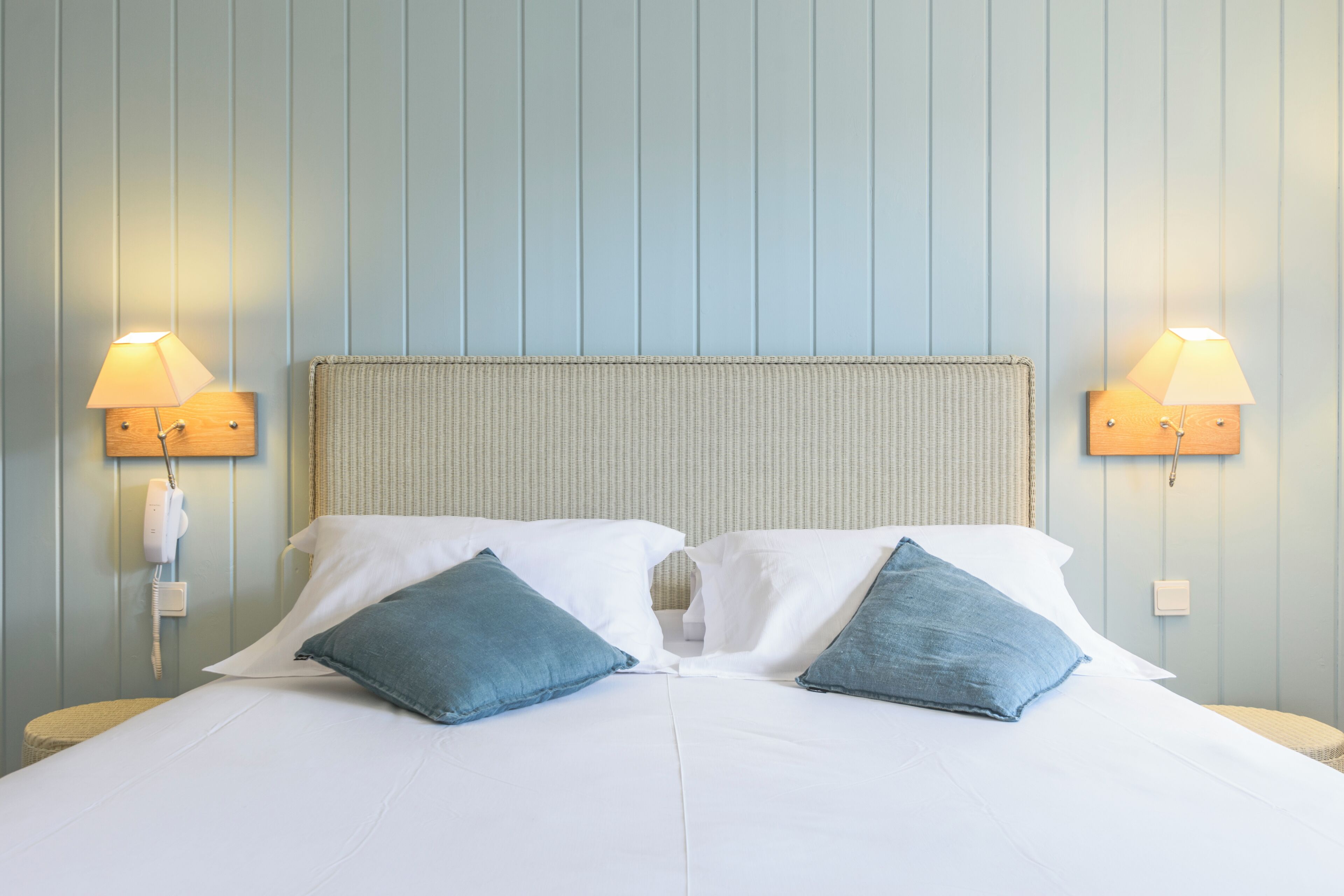 A neatly made bed with two pillows, a headboard, and two wall-mounted lamps on either side.