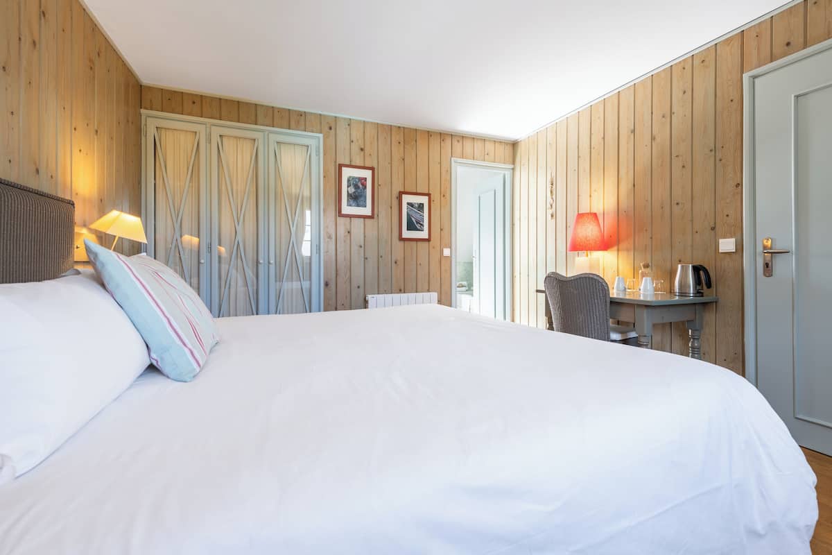 A bedroom with a large bed, wooden walls, a desk with a lamp, and a door.