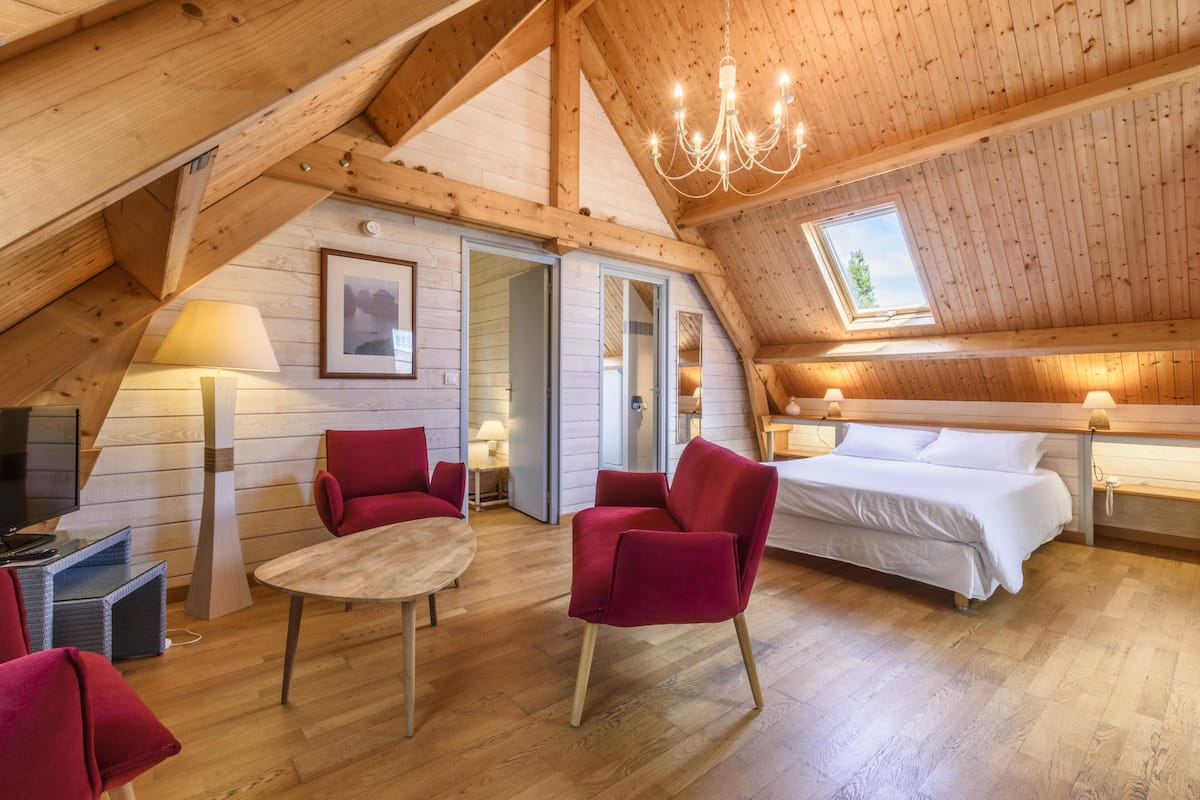 A cozy attic bedroom with a wooden ceiling, a chandelier, a bed, and a desk with a lamp.