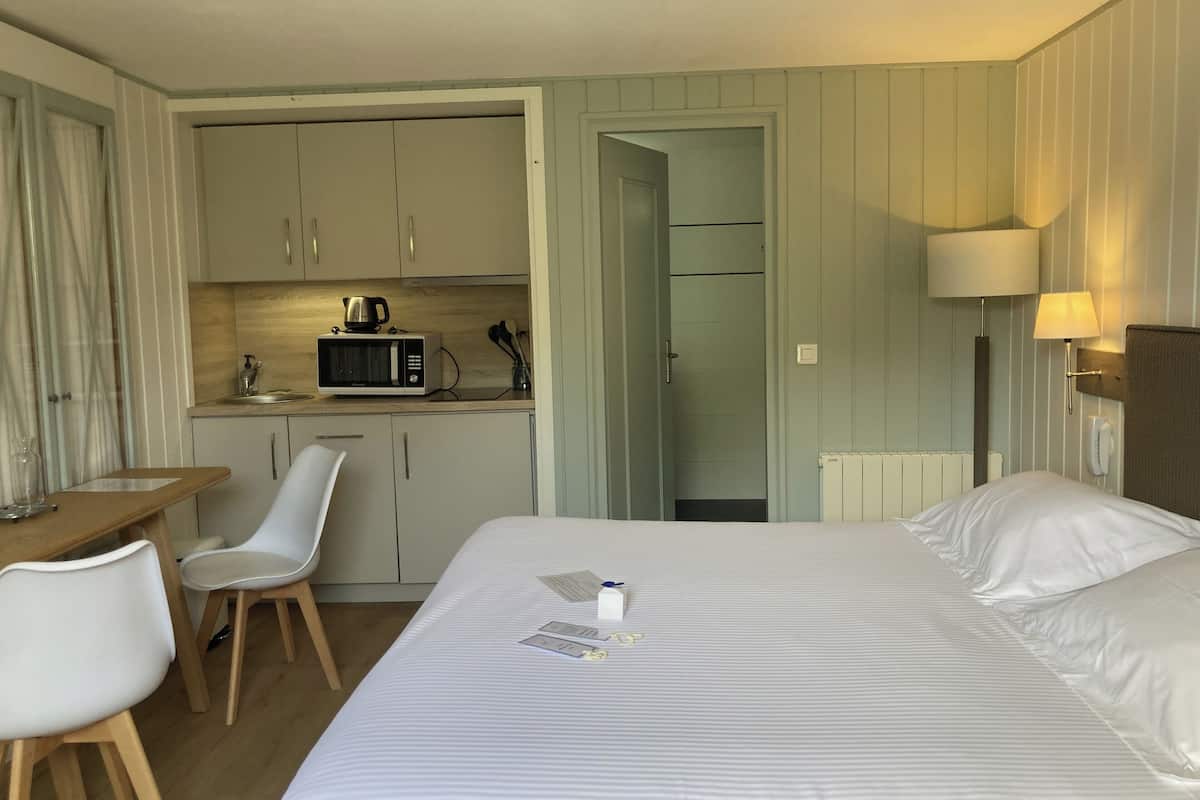 A compact hotel room with a bed, a desk, a chair, a kitchenette, and a bathroom.