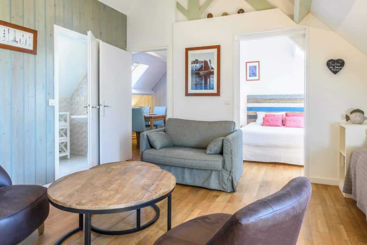 A cozy room with a wooden floor, a grey sofa, a round wooden table, and a bed with a striped headboard.