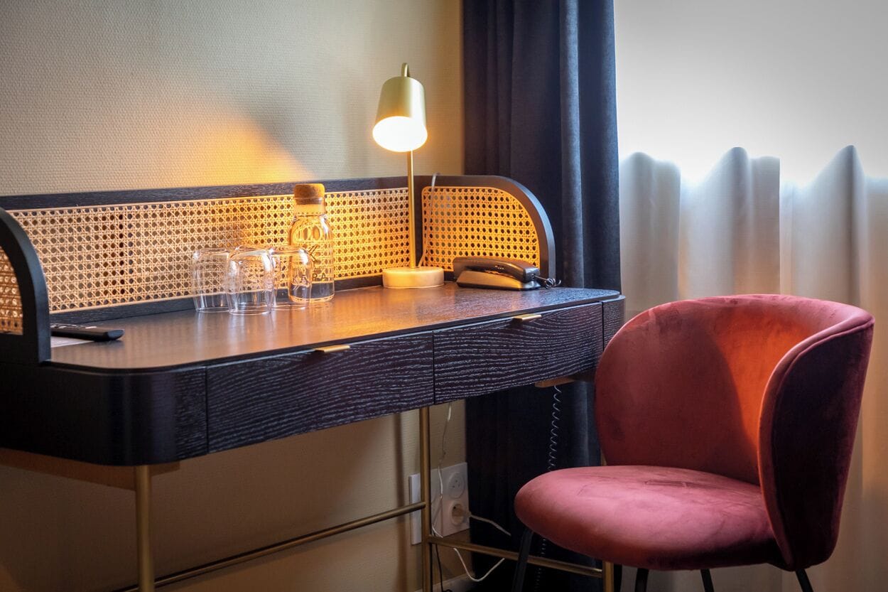 Una habitación de hotel con escritorio, lámpara, silla y ventana con cortinas.