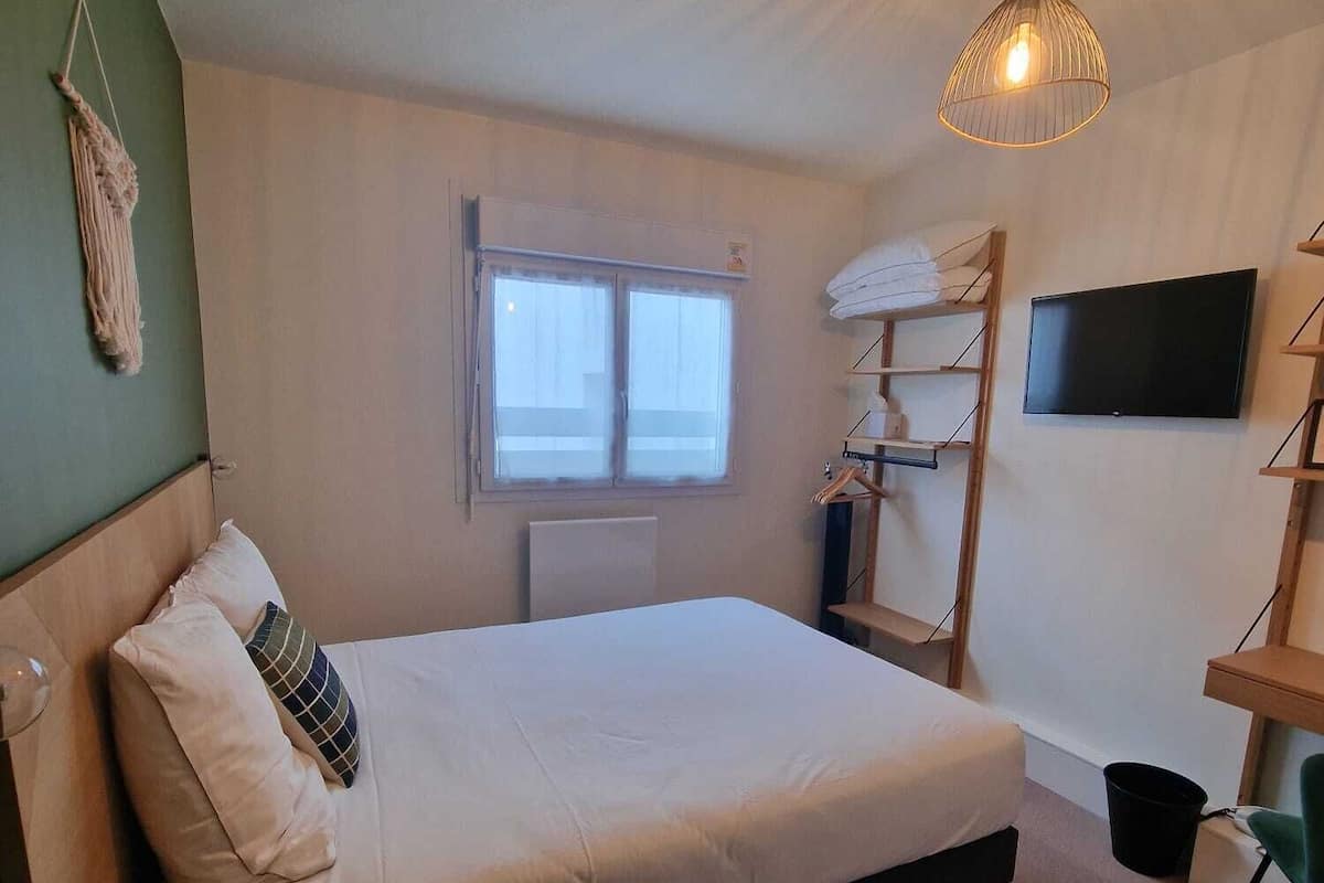 Un dormitorio con cama, escritorio, silla, televisor, ventana y lámpara colgante.