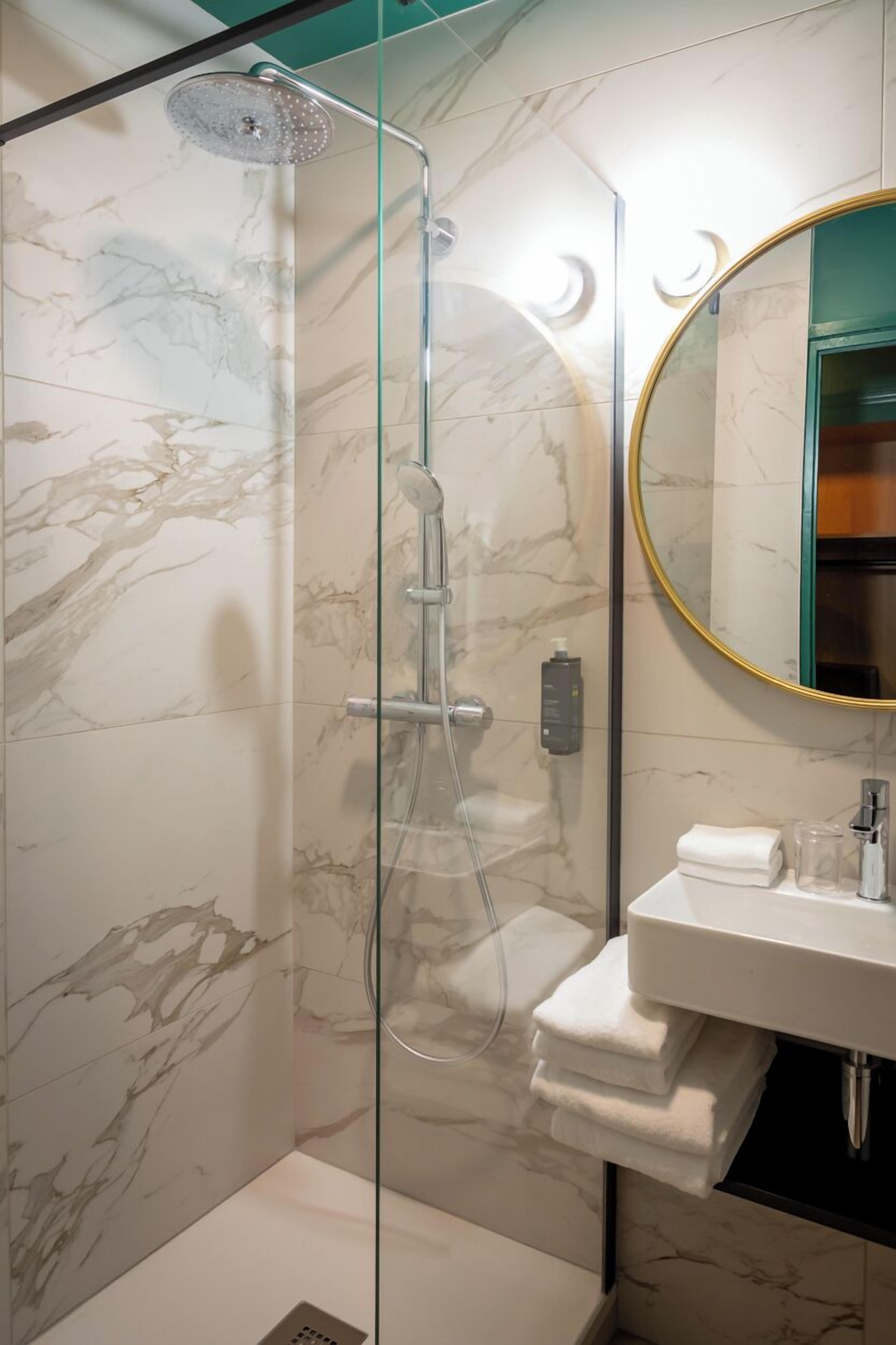 A modern bathroom with a glass-enclosed shower, marble walls, a round mirror, and a white sink with towels.