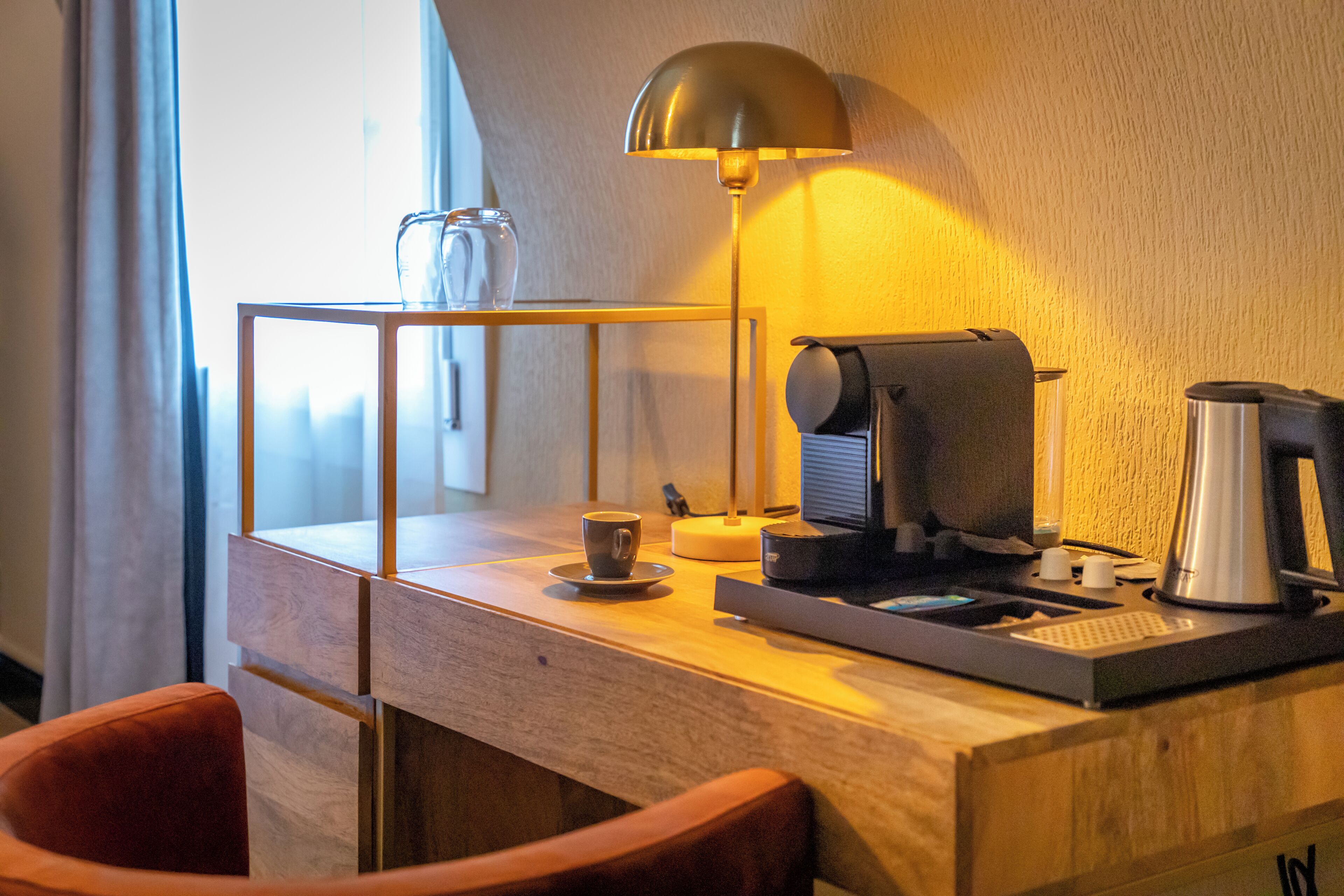 A hotel room with a wooden desk, a coffee maker, a lamp, a cup, and a chair.