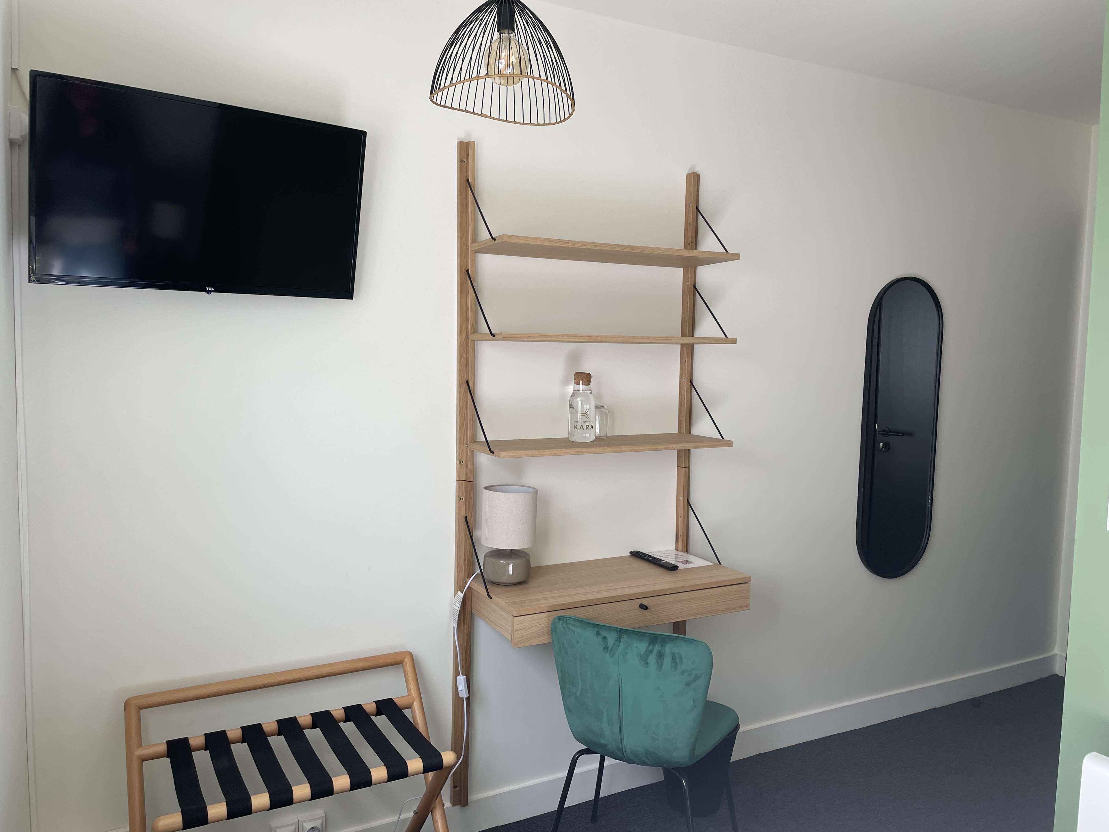 Une pièce dotée d’un téléviseur à écran plat fixé au mur, d’un bureau en bois avec une lampe, d’une chaise de couleur turquoise et d’une porte noire.