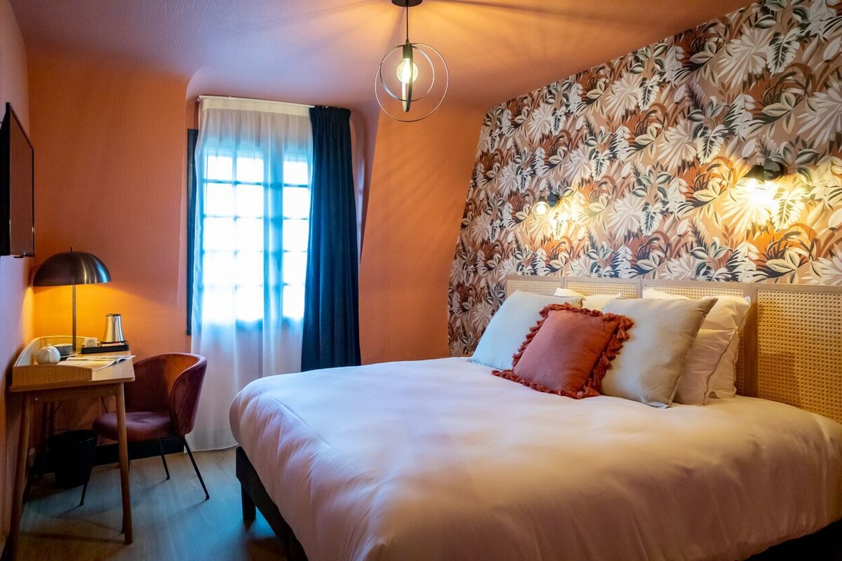 A hotel room with a bed, a desk, a chair, a lamp, and a window with curtains.