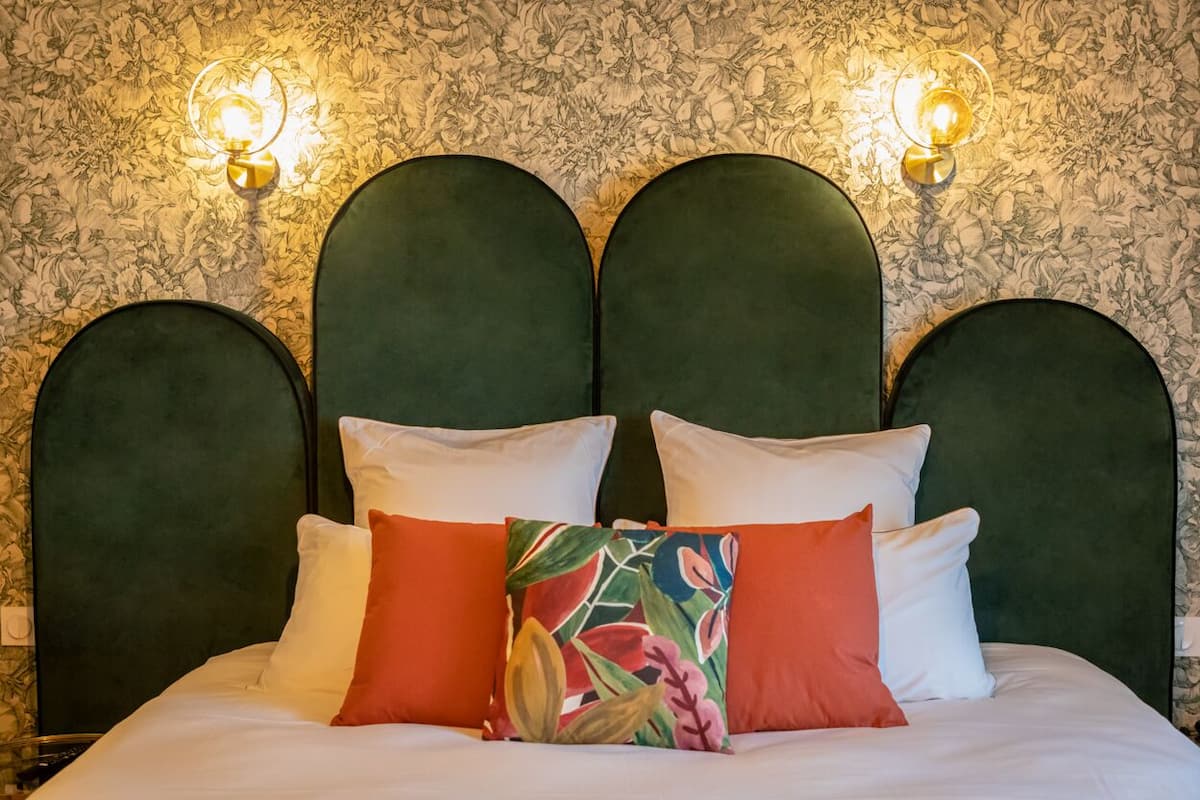 Una cama con cabecera tapizada de verde, almohadas blancas y anaranjadas, y papel tapiz con motivos florales.