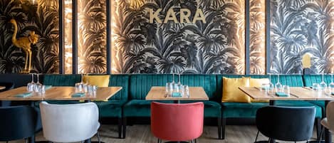 L’intérieur d’un restaurant présente un coin salon avec des banquettes recouvertes d’un tissu vert, des tables en bois avec des chaises, et un mur orné d’un motif de feuilles tropicales, avec le mot « KARA » inscrit au centre.