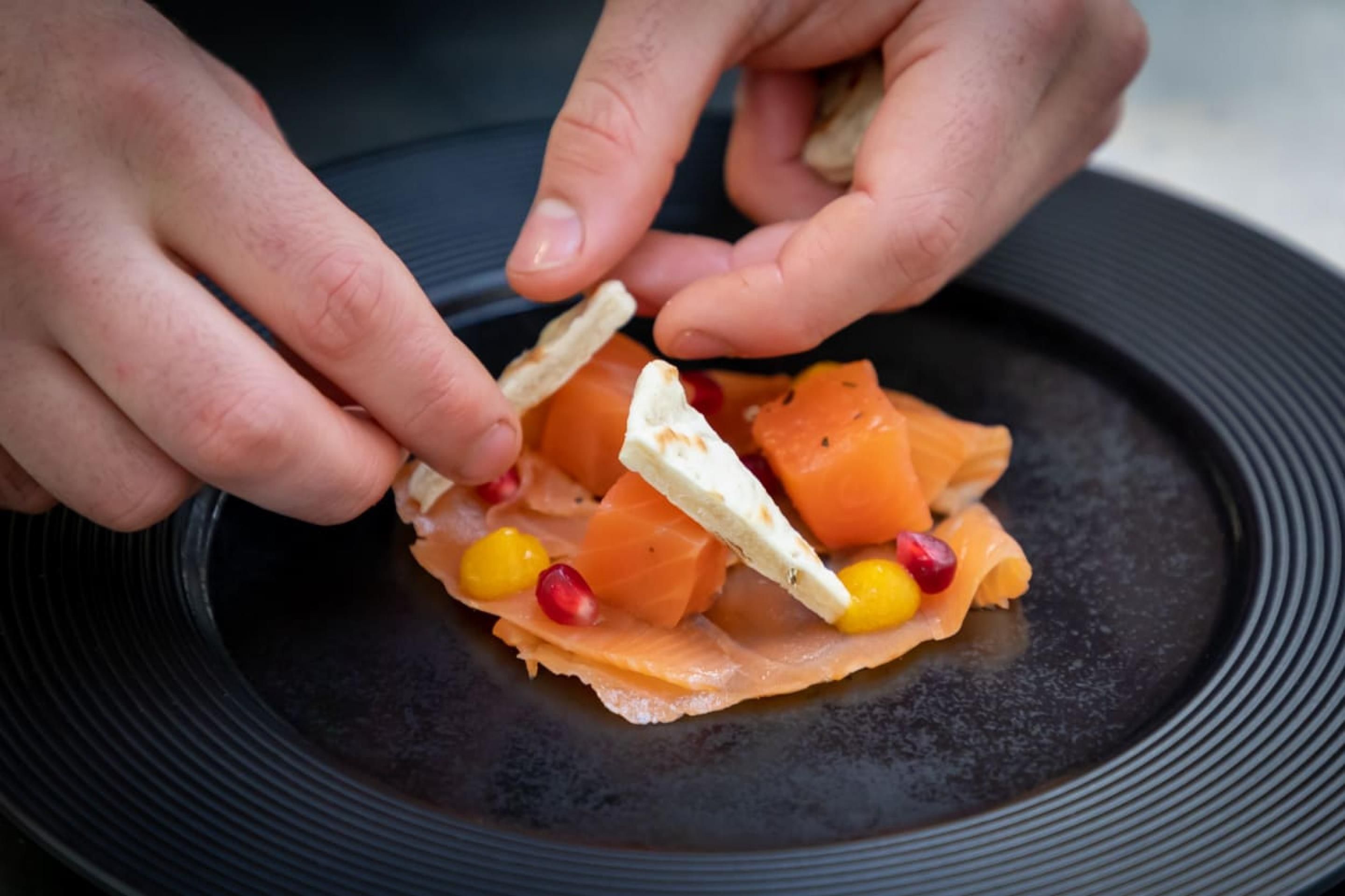 Eine Person legt einen Cracker auf einen Teller mit Räucherlachs, Pomegranate-Kernen und Mais.