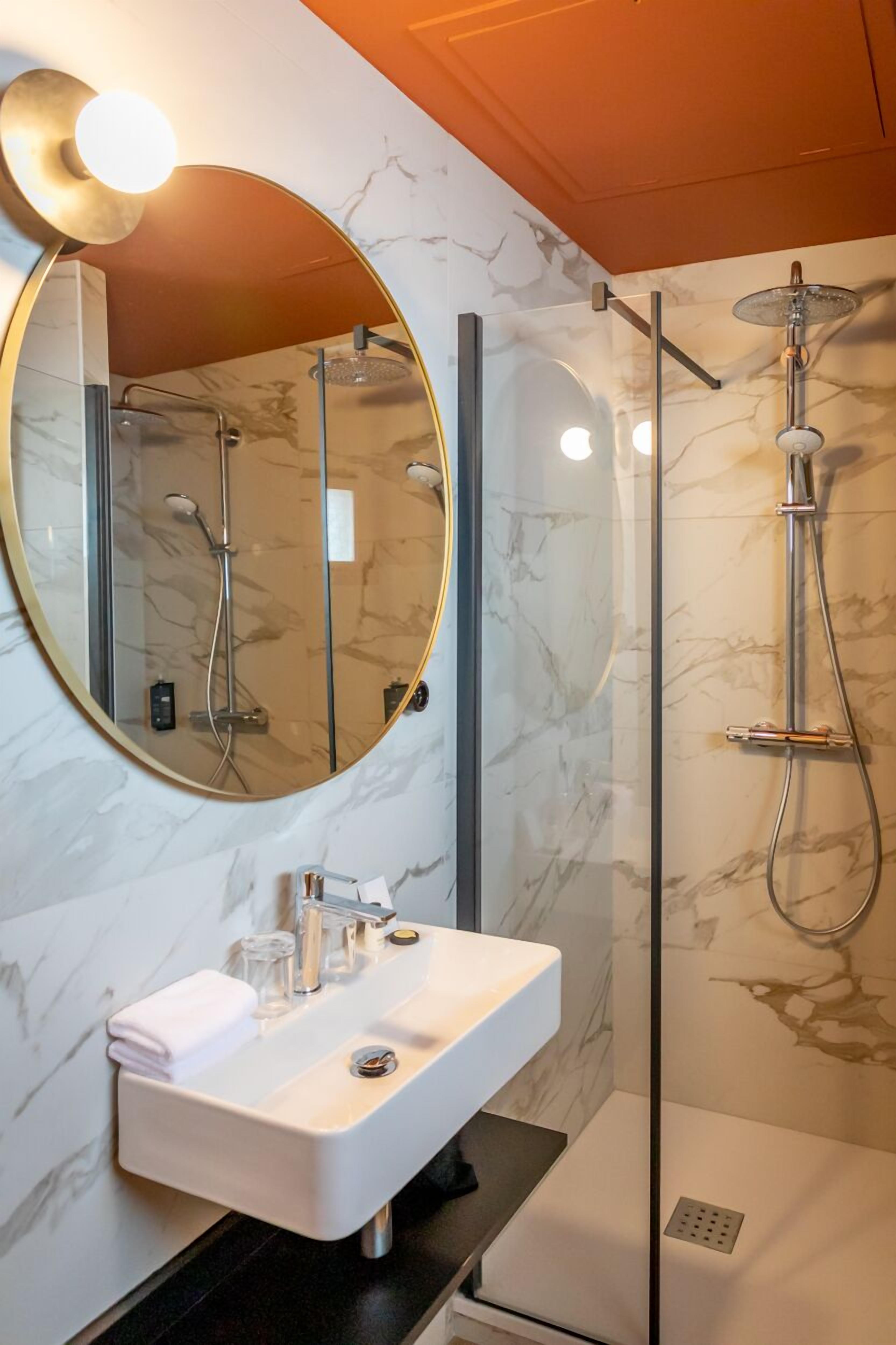 A modern bathroom with a marble wall, a round mirror, a white sink, and a glass-enclosed shower.