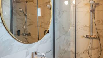 A modern bathroom with a marble wall, a round mirror, a white sink, and a glass-enclosed shower.