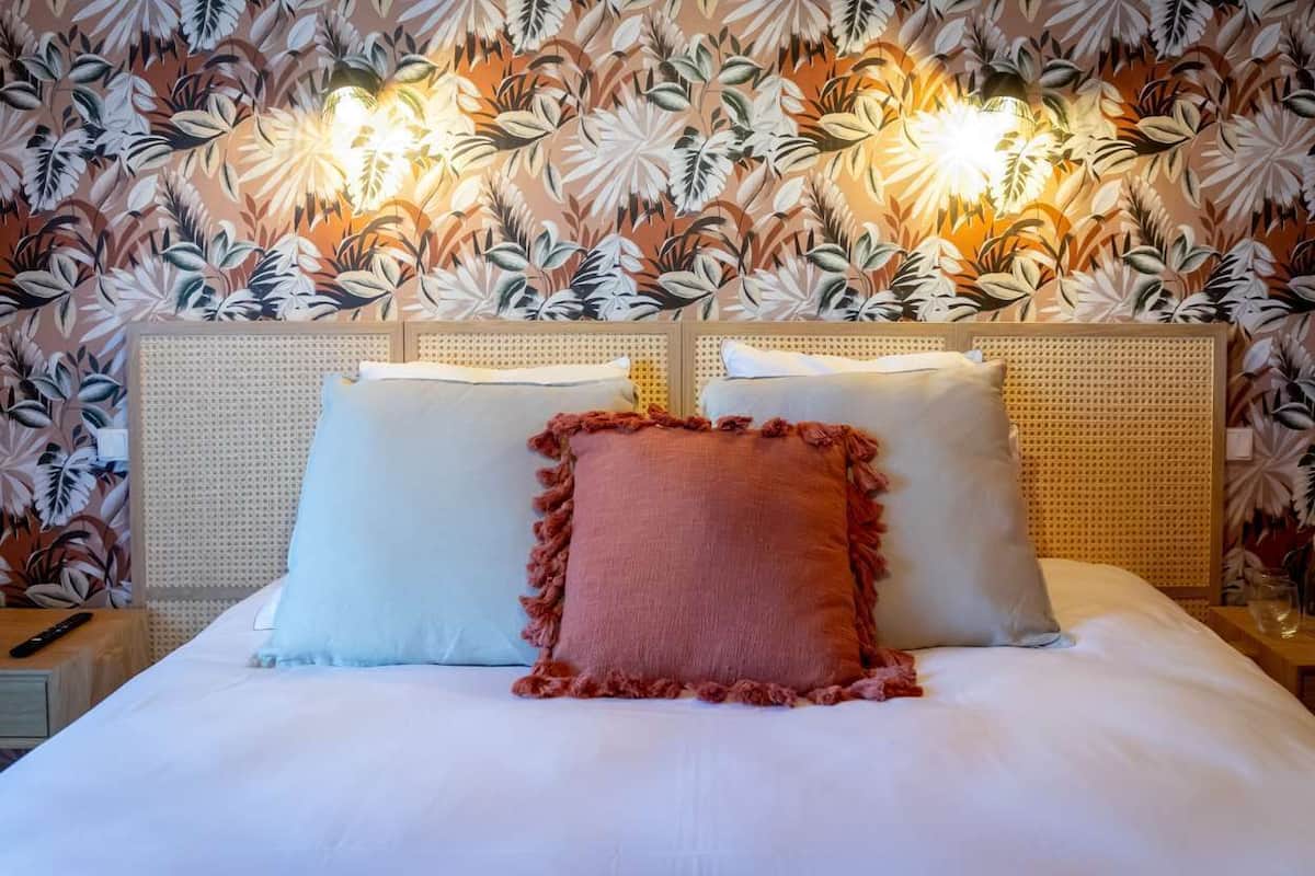 Una cama con dos almohadas, una pared con estampado floral y una mesita de noche con un control remoto.
