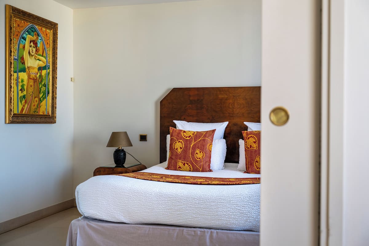 A neatly made bed with white linens and patterned pillows, a wooden headboard, a nightstand with a lamp, and a framed painting on the wall.