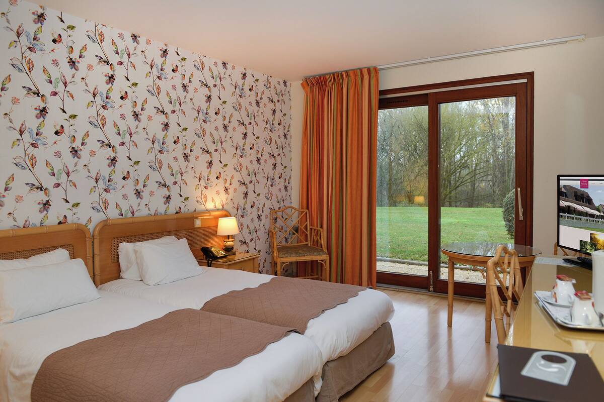 A hotel room with a large bed, a desk with a computer, a chair, and a view of a garden through a sliding glass door.