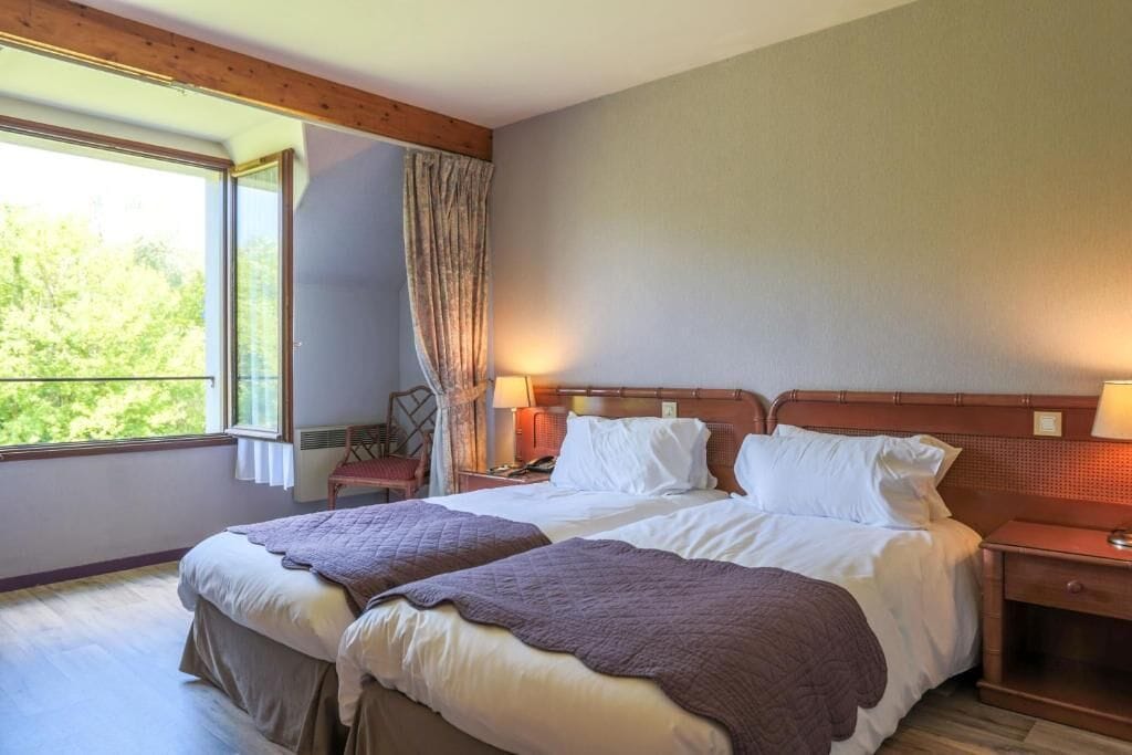 Una cama doble con cabecera, dos mesitas de noche, una lámpara de noche, una ventana con cortinas y vistas al exterior.